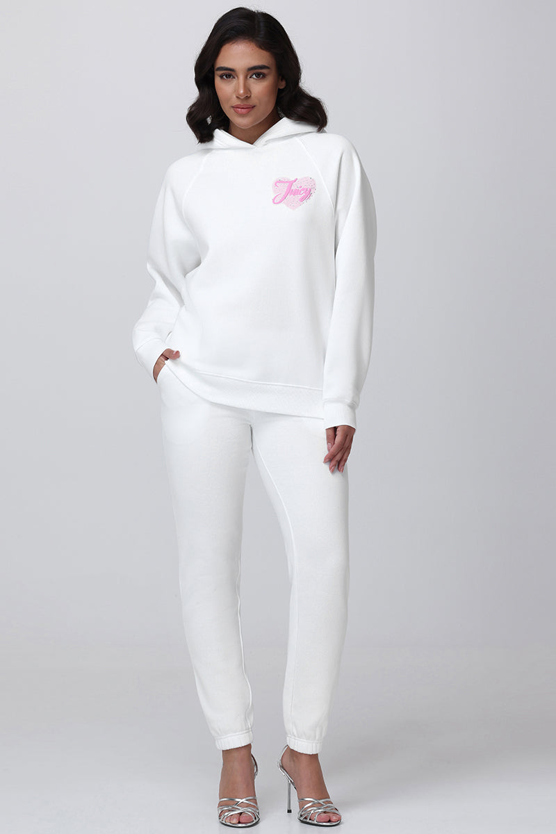 A woman with dark hair stands against a plain background wearing Sweetheart Fleece Sweatpants and a white hoodie with pink Juicy lettering, paired with silver open-toe heels.
