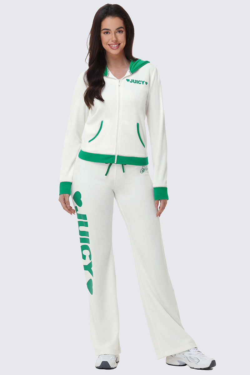 A woman with long dark hair stands against a light gray background, wearing Contrast Low Rise Track Pants—a sporty white tracksuit featuring green trim and bold "JUICY" lettering on the left leg.