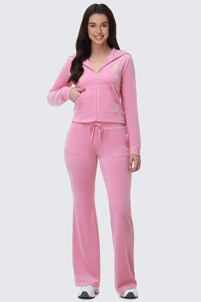 A woman smiles, wearing Heritage Cotton Velour Track Pants in pink with a zip-up hoodie and drawstring waist, paired with white sneakers, against a light gray background.