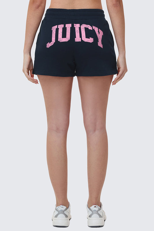 A person is seen from behind wearing Collegiate Booty Shorts in black terry with a bold pink Juicy logo across the back, paired with white sneakers.