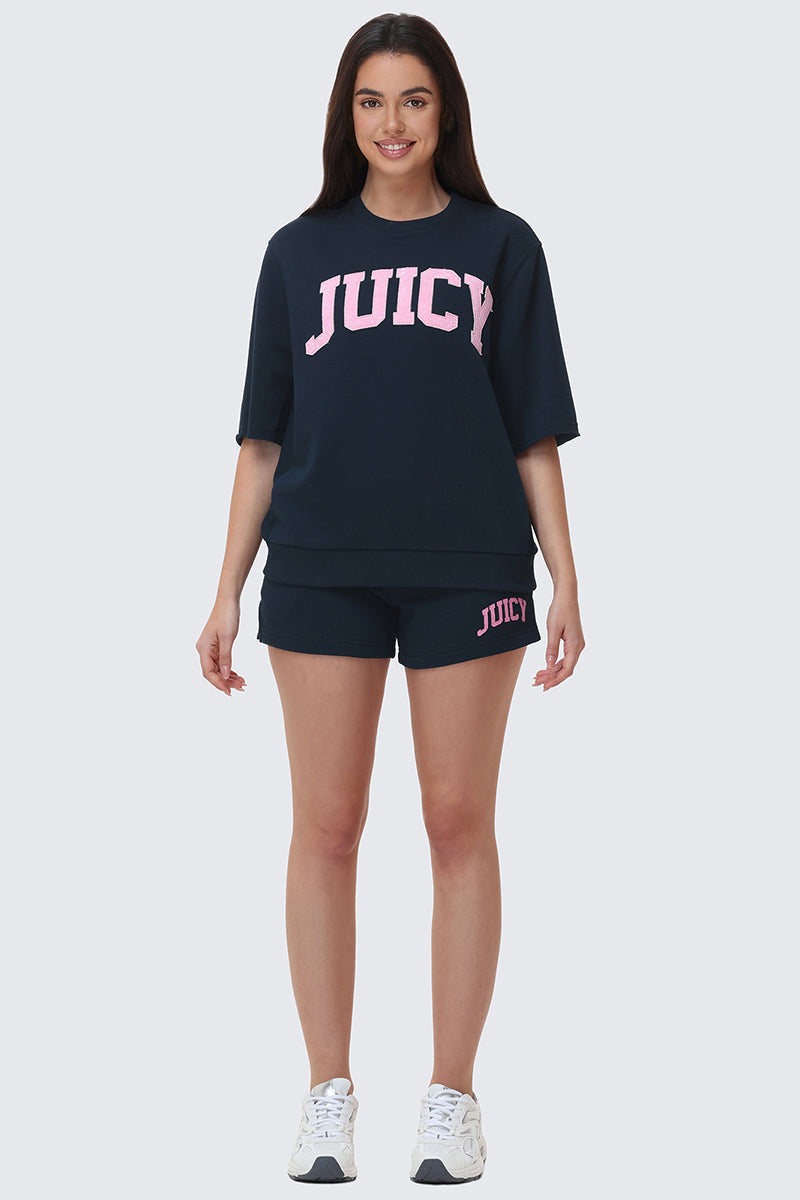 A woman smiles facing forward, wearing navy Collegiate Booty Shorts with a drawstring waist and pink Juicy logo, a matching top with a large pink Juicy logo, and white sneakers.