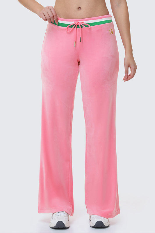 Someone models the Heritage Stripe Bling Velour Track Pants—pink wide-leg velour with a green and white Juicy-logo waistband—paired with white sneakers. Only the lower torso and legs appear against a plain background.