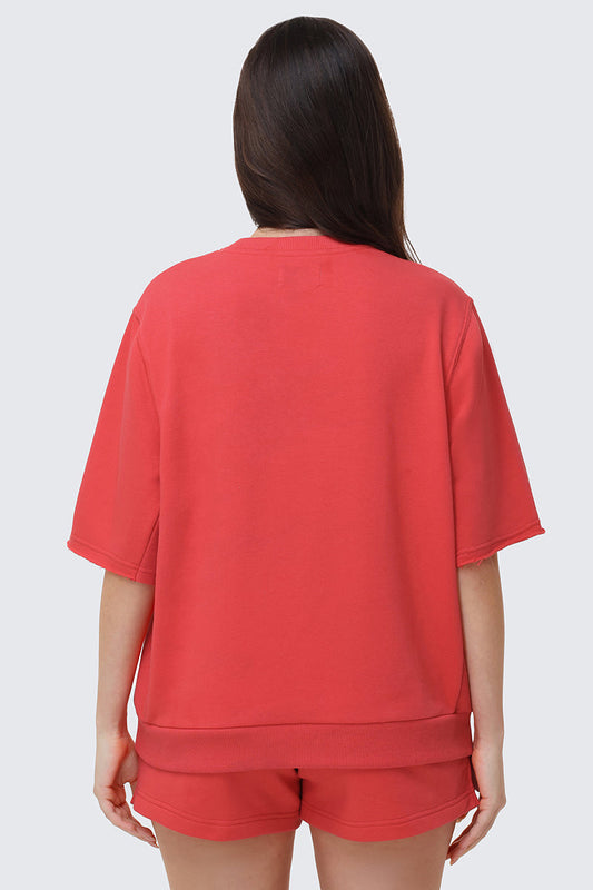 A person with long dark hair stands with their back to the camera, wearing a relaxed fit red Collegiate Sweatshirt and matching shorts, evoking a collegiate-style look against a plain light background.