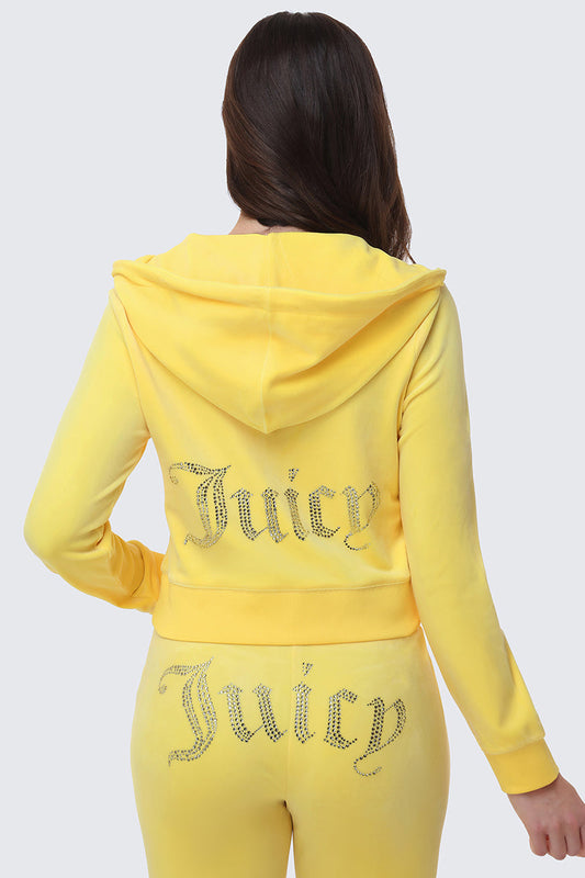A woman with long brown hair, facing away from the camera, wears the OG Big Bling Velour Hoodie—a yellow tracksuit with a hood and luxe rhinestone-studded Juicy text embellishment on the back of both jacket and pants.