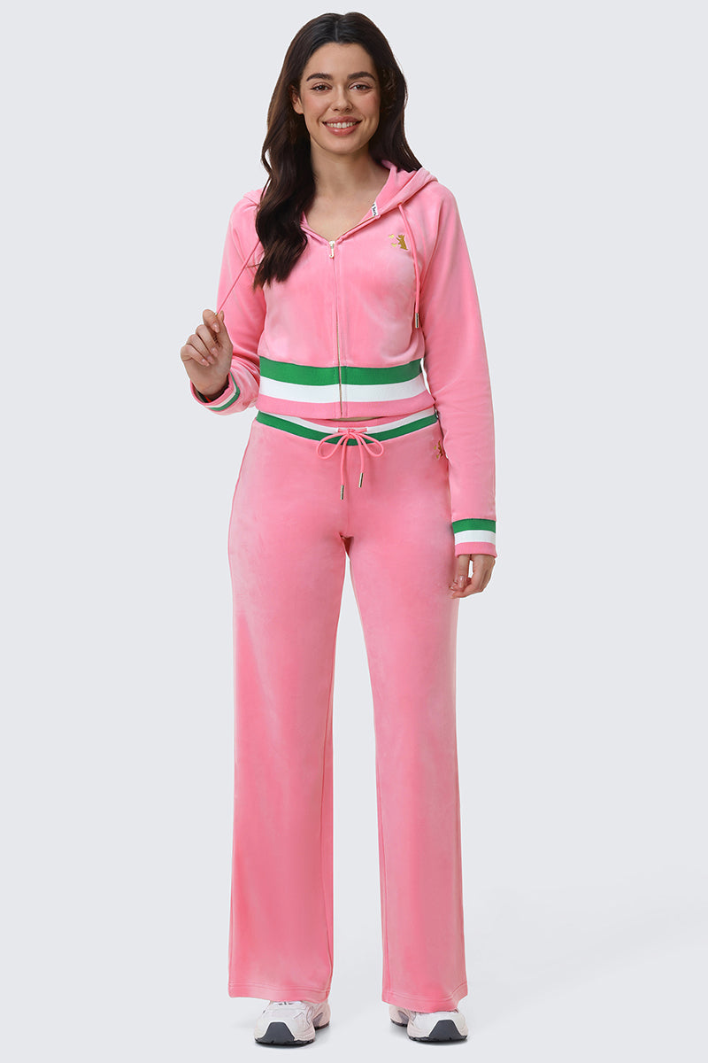 A woman smiles in the Juicy Couture Heritage Stripe Bling Velour Zip Hoodie and matching drawstring pants with green and white striped bands, rhinestone logo, and white sneakers, against a plain light gray background.