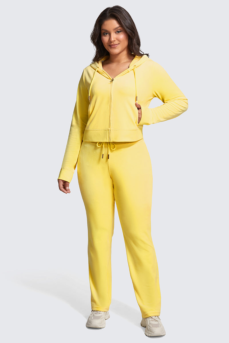 A woman smiles and faces forward, wearing the Plus-Size OG Big Bling Velour Hoodie in yellow with matching sweatpants and white sneakers. The hoodie features a front zipper and drawstring, adding a luxe touch. Her left hand rests in her pocket.