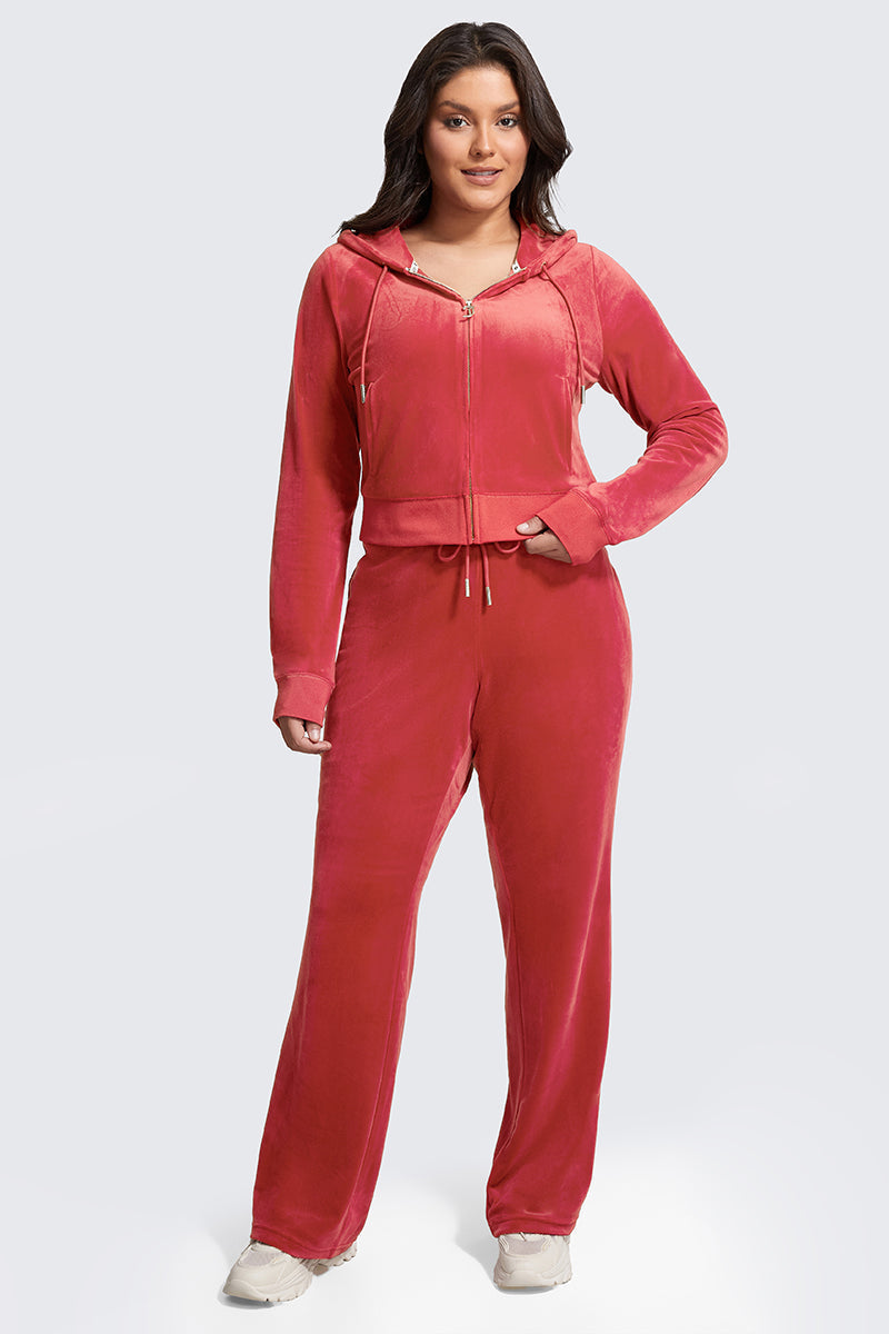 A woman with long dark hair smiles slightly while facing forward, wearing the Plus-Size OG Big Bling Velour Hoodie in pink with matching drawstring pants and white sneakers, set against a light gray background.