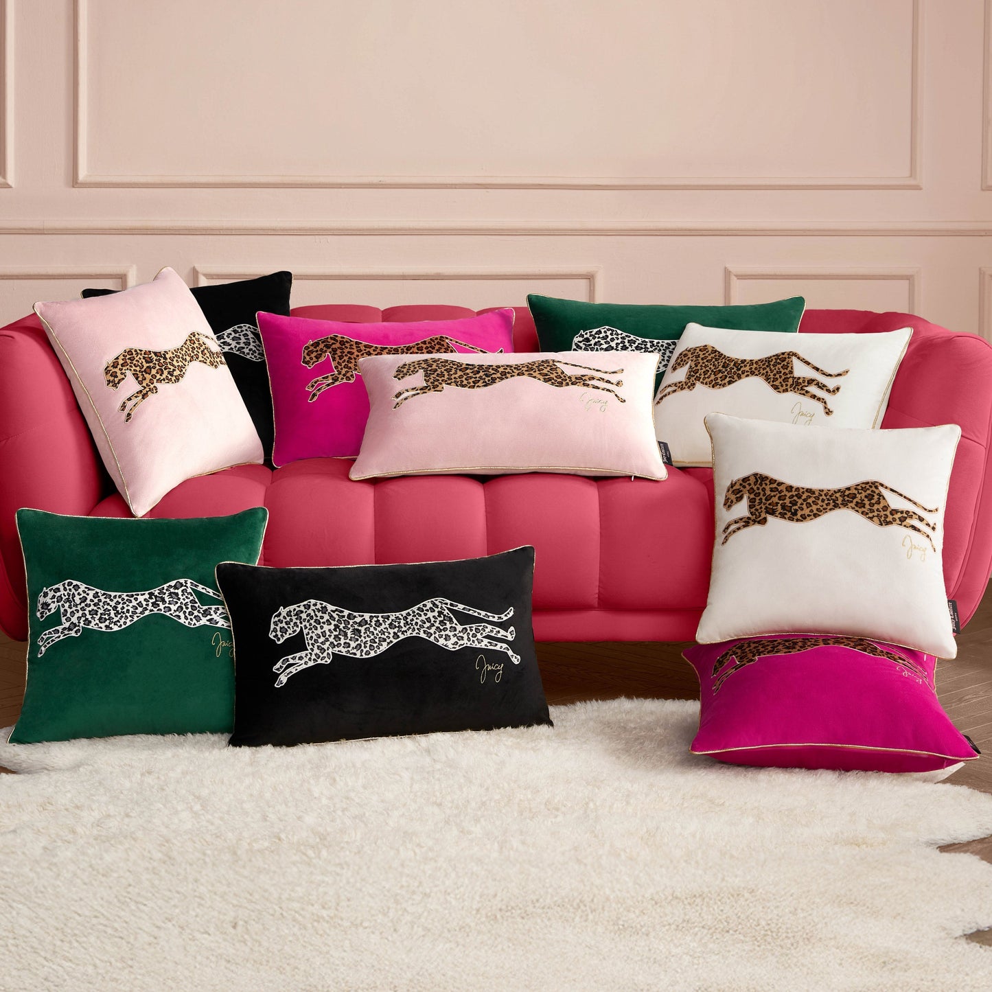 A pink sofa with assorted pillows, including the Velvet Cheetah Pillow and others in black, white, green, and hot pink covers, is set against a pale pink wall. A white fluffy rug lies on the floor.