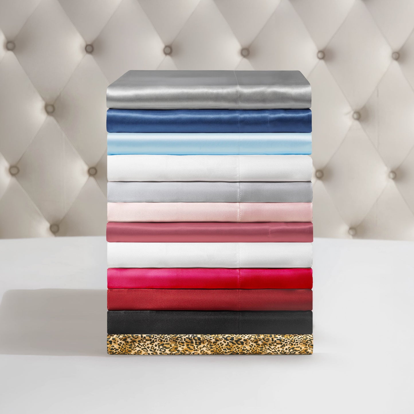 A neatly stacked pile of Solid Satin Sheet Sets in assorted colors and patterns—grey, blue, white, pink, red, black, and leopard print—displayed against a tufted beige background.