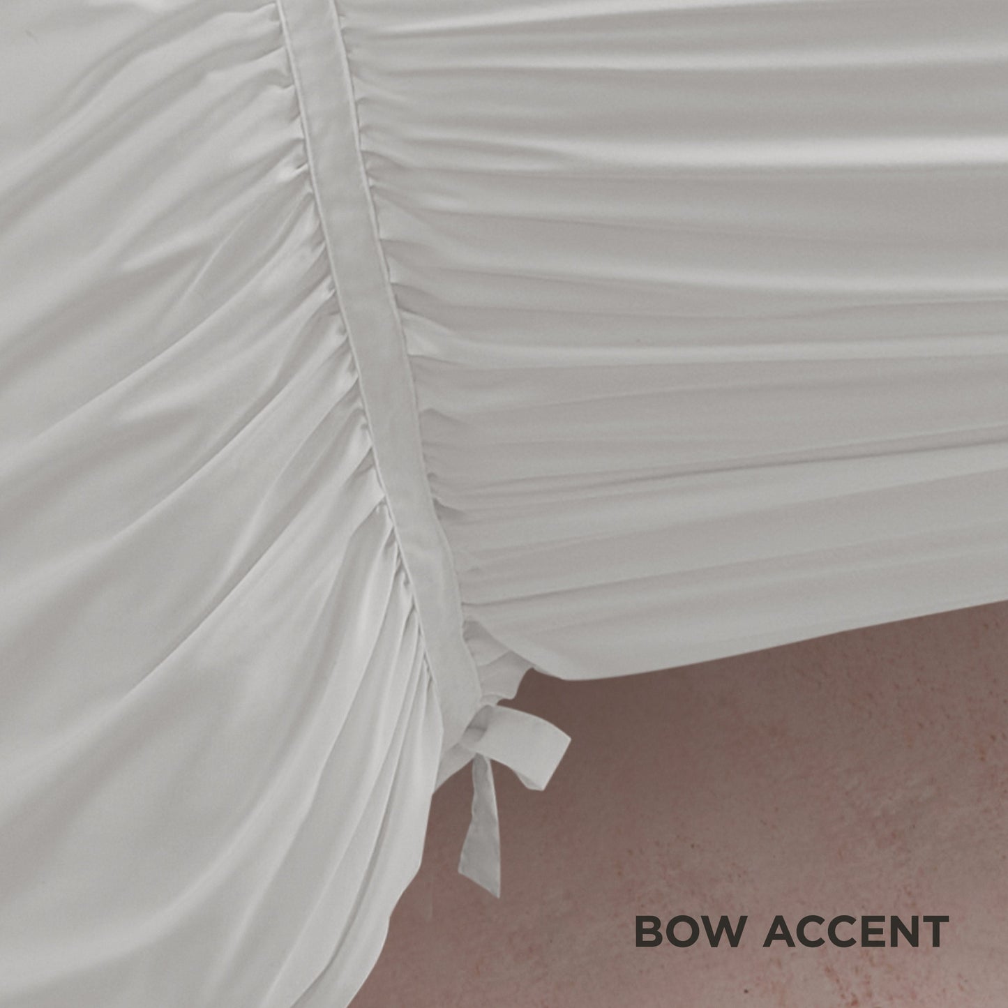 Close-up of the Ribbon Ruched Comforter Set in white, featuring ribbon detailing and a side bow accent, draped over an edge with a pink floor beneath. "BOW ACCENT" appears in black text at the lower right corner.