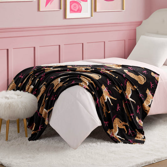 A neatly made bed with a white comforter and a Plush Throw Blanket featuring cheetah and pink bow patterns. The room has pink walls with framed art, a white furry stool, and a white rug on the floor.
