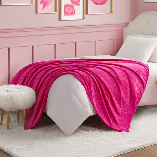 A neatly made bed features a Plush Throw Blanket, bright pink and ultra-soft, draped over white bedding in a pink-themed room. A fur-topped stool and framed wall art complete the cozy, stylish decor.