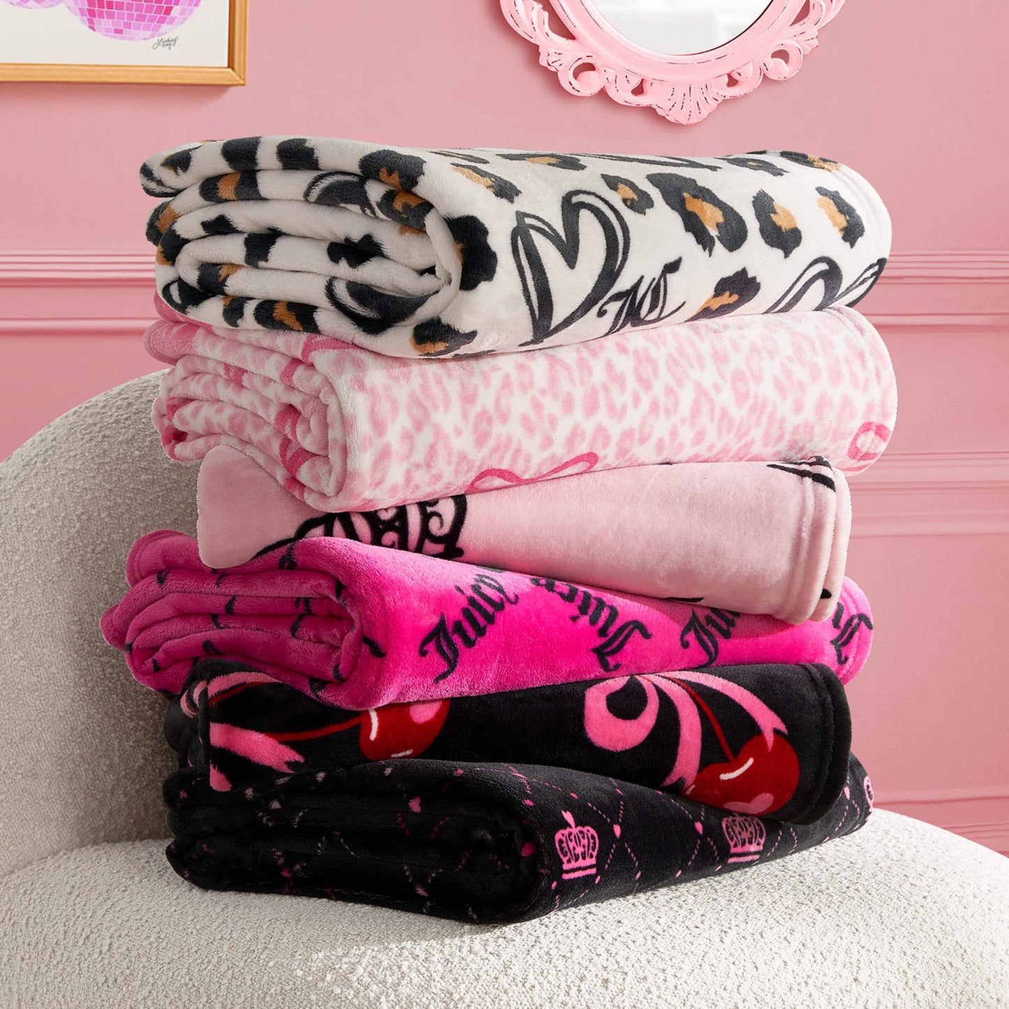 A stack of five folded Plush Throw Blankets in assorted pink, black, and animal print patterns rests on a white textured chair beneath decorative frames on a pink wall.