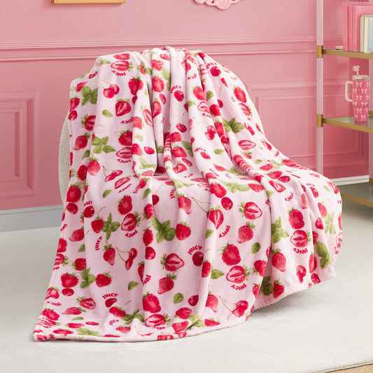 The Plush Throw Blanket features soft pink fleece with red strawberries, green leaves, and “JUICY” lettering—perfect for draping over a chair to accent a pink-themed room with a gold-and-glass shelf in the background.