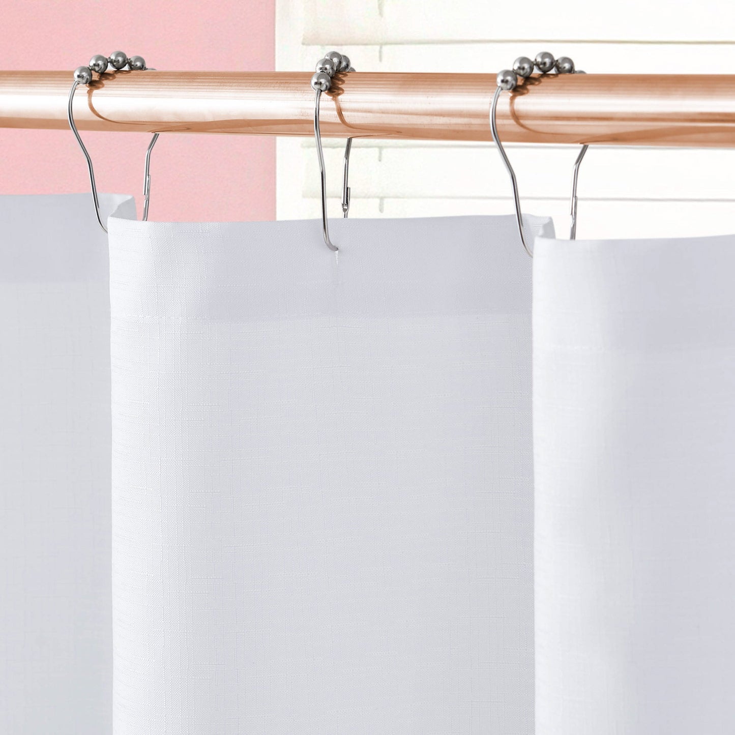 Close-up of the Shower Curtain and Hooks Set hanging from a light wooden rod with silver hooks, featuring ball bearings, against a pink wall and white blinds for a chic, modern look.