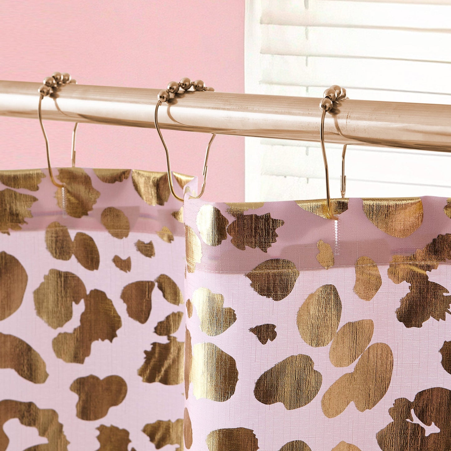 Close-up of the Shower Curtain and Hooks Set featuring a gold and pink leopard print, hanging from a silver rod with metal rings. The background shows stylish bathroom decor, including a pink wall and partially open white blind.
