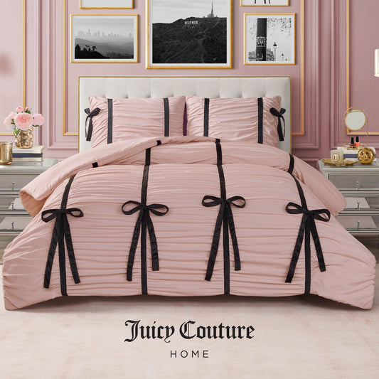 A blush pink Amelie Bows Ruched Comforter Set by Juicy Couture, featuring black velvet bows, styled in an elegant bedroom with pink walls, gold-framed art, and a white dresser. Ultra-soft comforter with Juicy Couture Home logo at the bottom.