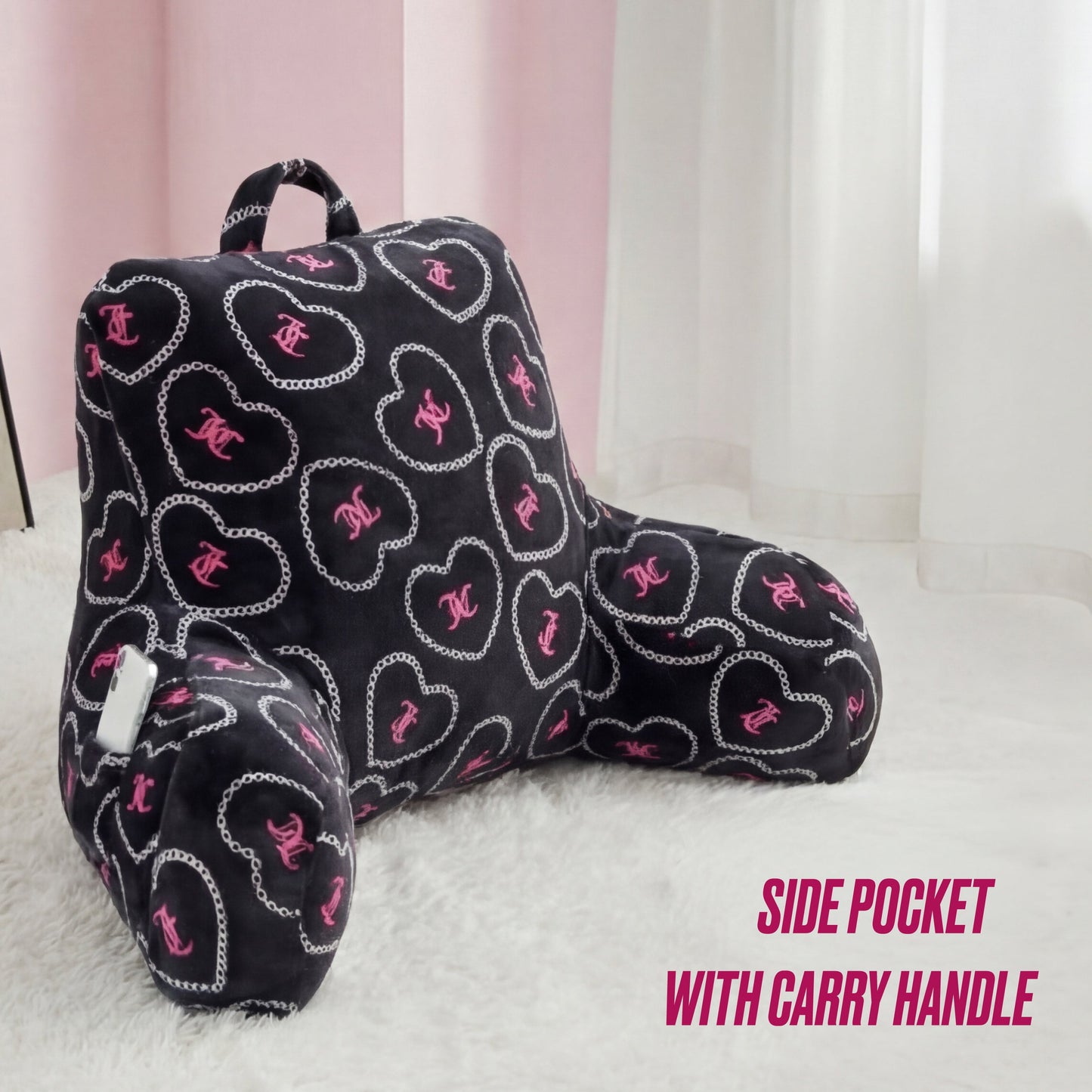 A Juicy Couture Backrest Pillow in pink and white with heart and chain patterns sits plush on the floor near sheer curtains, featuring a side pocket and carry handle. Text: SIDE POCKET WITH CARRY HANDLE.