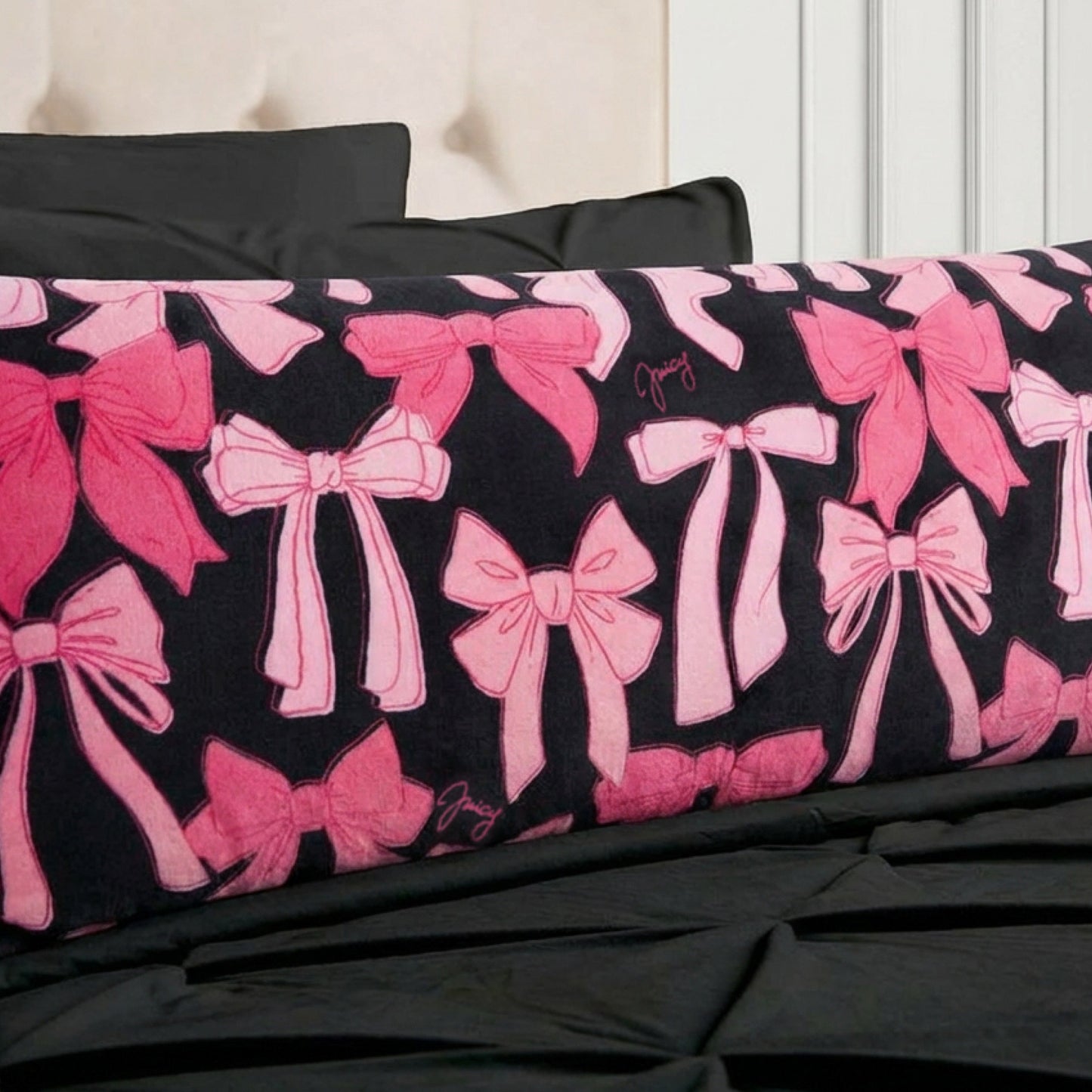 The Plush Body Pillow by Juicy Couture features pink bows and cursive signatures, adding comfort and designer flair to your space. Styled on a bed with black bedding and a beige tufted headboard for a chic look.