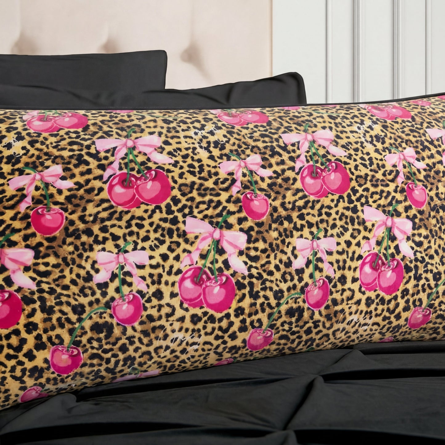 The Plush Body Pillow features a leopard print background with bright pink cherries tied with light pink bows, and is shown resting on a black bed with matching black pillows.