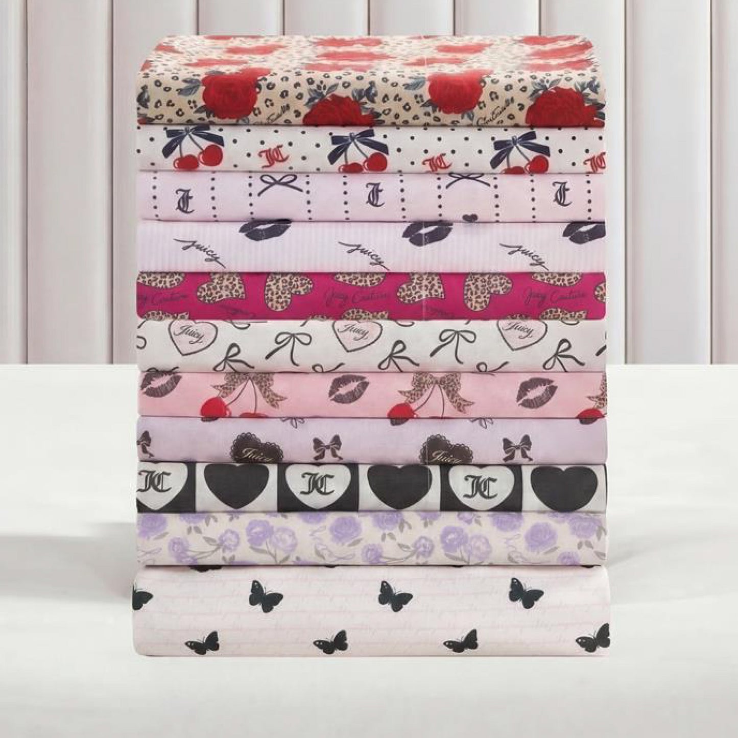 Eight Printed Microfiber Sheet Sets with colorful patterns—including roses, cherries, bows, hearts, lips, animal print, and butterflies—are neatly stacked against a light vertical-striped background for a luxurious bedding style.