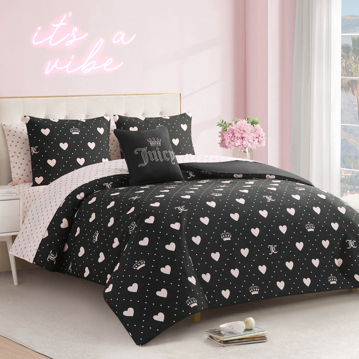 A neatly made bed with the Diamond Hearts Black Comforter Bed in a Bag Bedding Set, featuring white hearts and crowns. Glam pillows include one that says Juicy. The room has pink walls, a neon "it's a vibe" sign, and a window view.