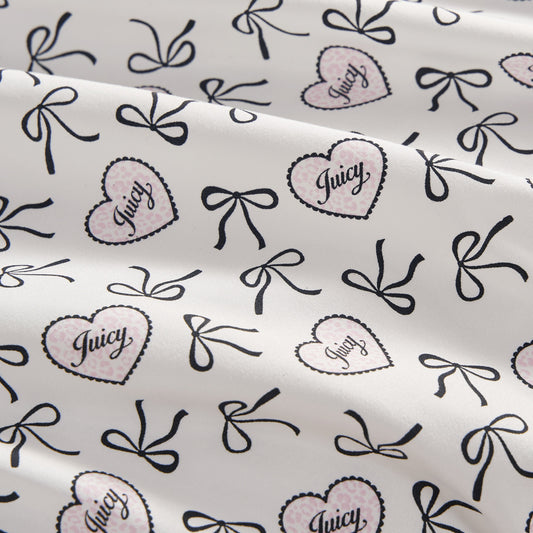 The Printed Microfiber Sheet Set features a white base with black bows and pink polka-dotted hearts outlined in black, each heart labeled "Juicy" in script. The luxury bedding is softly draped with gentle folds.