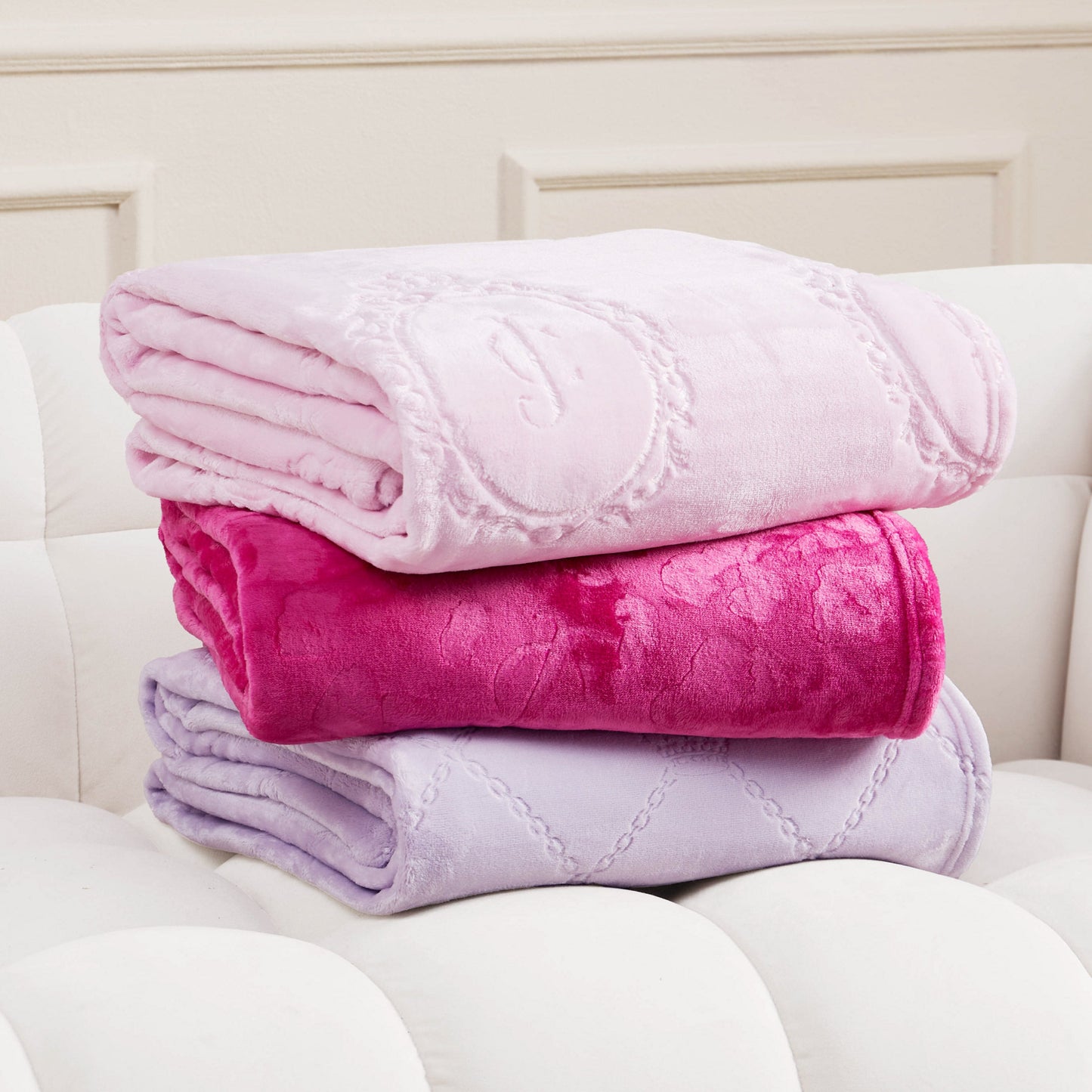 Three folded Juicy Couture Plush Oversized Throw Blankets, 60" x 90", in pastel pink, bright pink, and lavender with a subtle embossed pattern are stacked on a white tufted sofa.