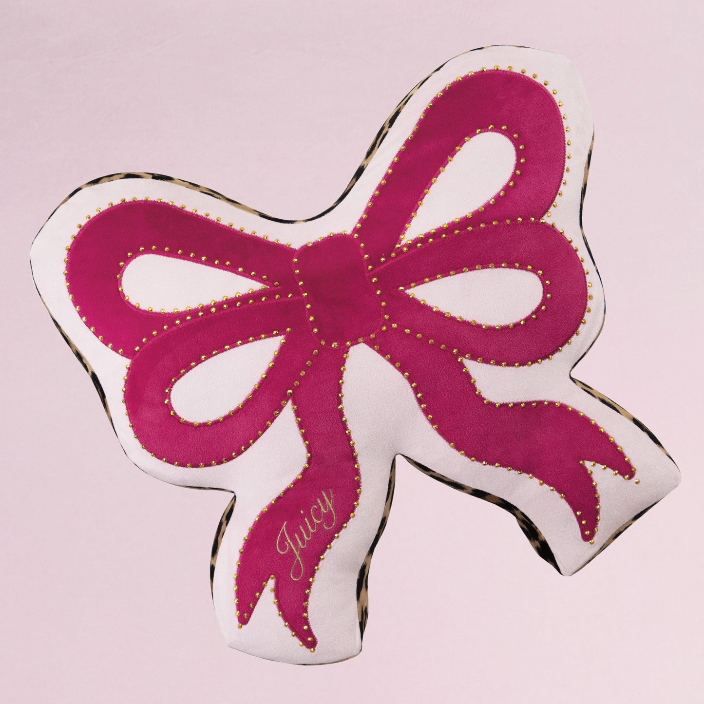 The Reversible Bow Decorative Pillow by Juicy Couture features pink and white fabric, gold stitching, and “Juicy” embroidered on one tail. Its bow shape adds a stylish touch to any space, set against a light pink background.