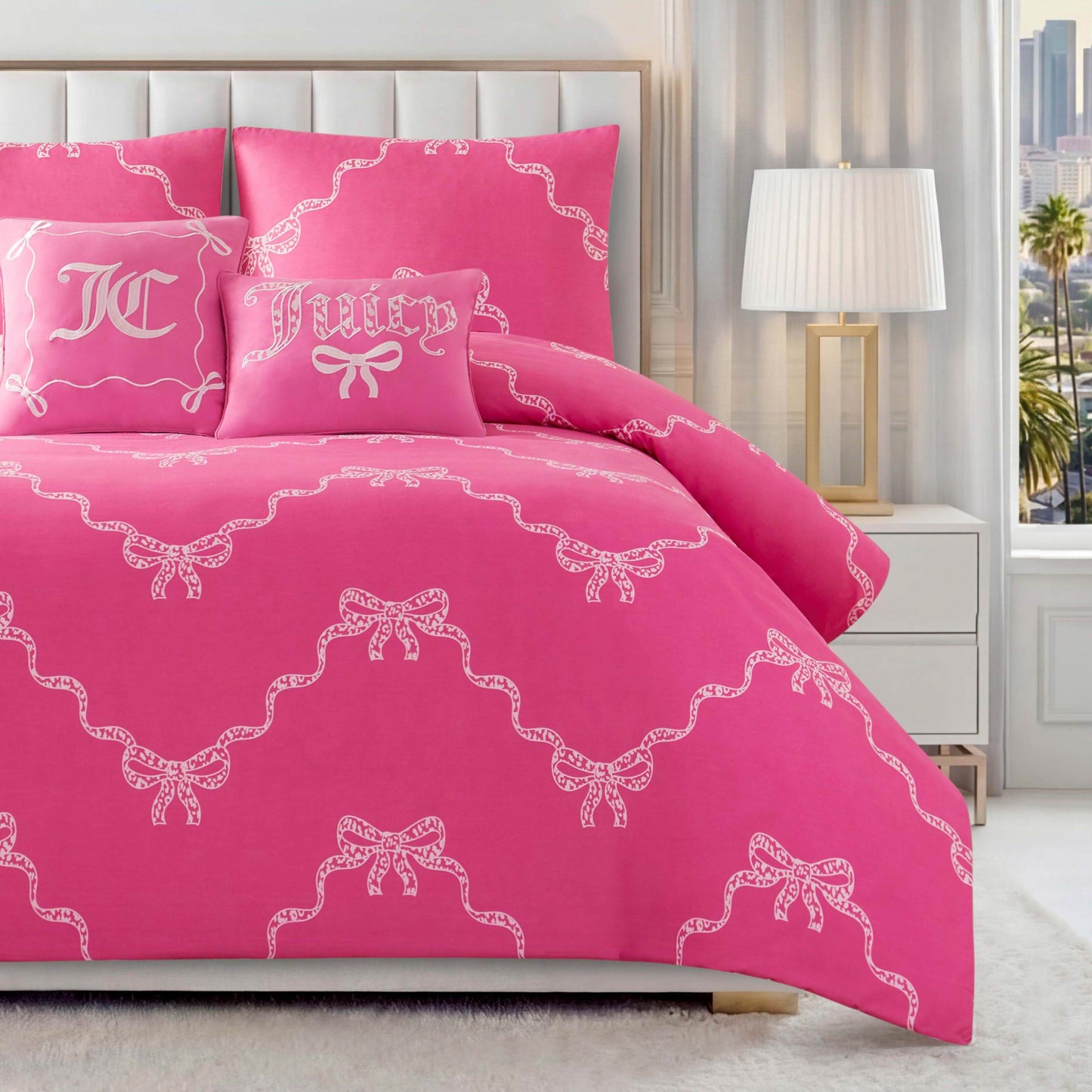 The Chevron Leopard Bow Reversible Comforter Set—pink with white bows and lace, plus embroidered pillows featuring JC initials and "Juicy" text—adds a playful pop to a modern bedroom with white furniture and large window.