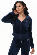 A woman wearing a Custom Initial OG Big Bling Velour Hoodie in navy blue, paired with matching drawstring pants and a rhinestone-embellished zip-up jacket, stands looking to the side with one hand touching her hair.
