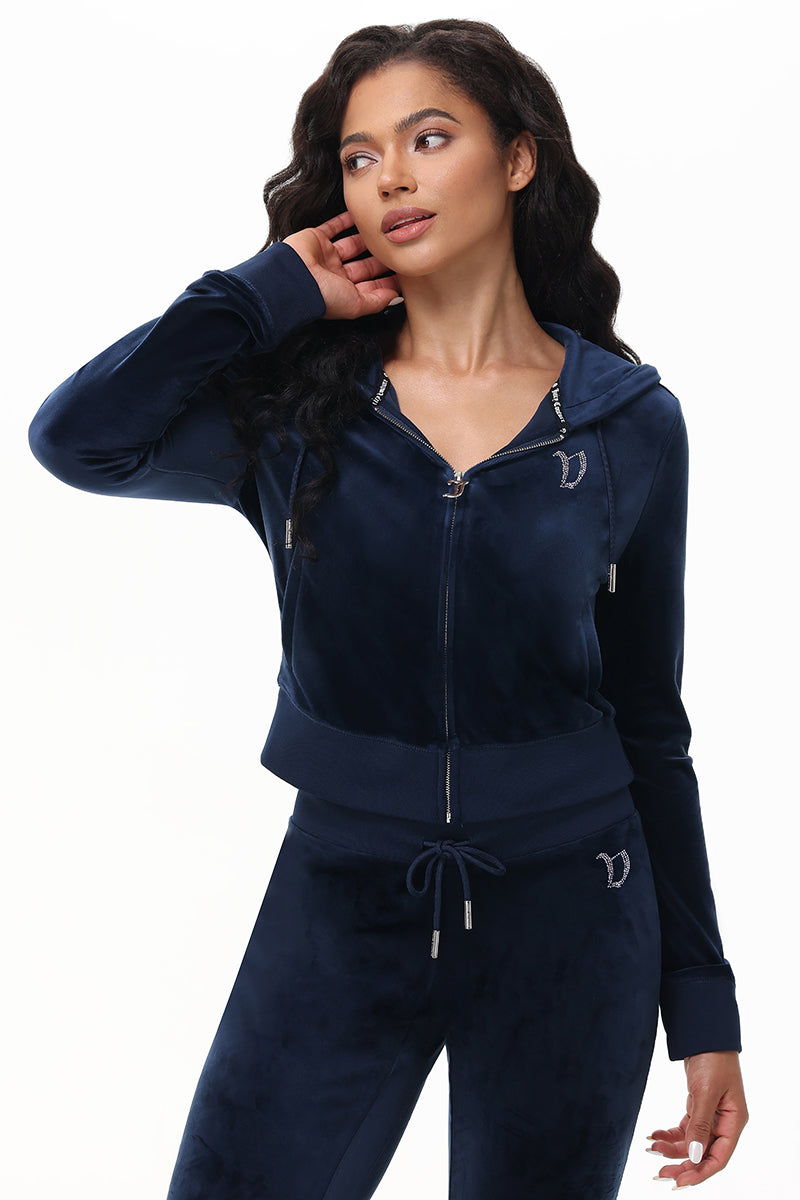A woman wears the Custom Initial OG Big Bling Velour Hoodie in dark blue, featuring metallic accents, Juicy rhinestones, a hood, and decorative emblems on the chest and hip as she looks to the side with one hand in her hair.
