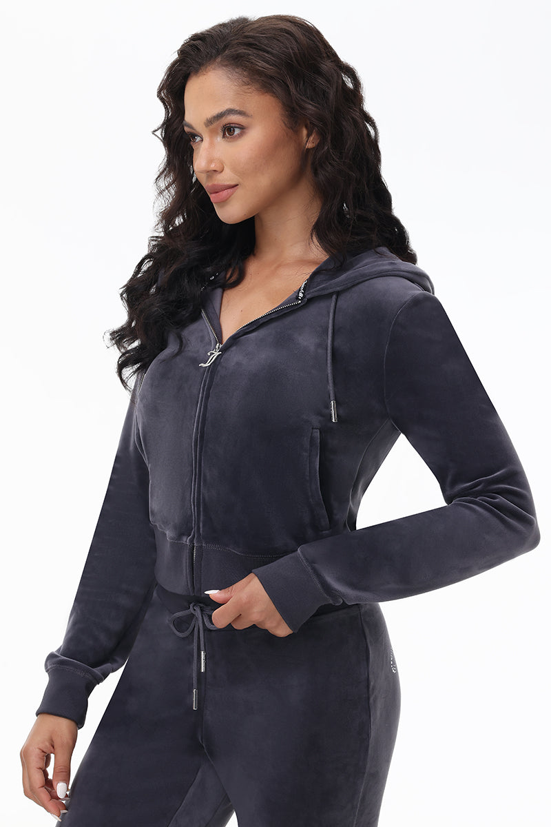 A woman models the OG Big Bling Velour Hoodie in dark gray, featuring thumbholes and a zip-up front, paired with matching pants, standing with one hand on her waist against a plain white background.