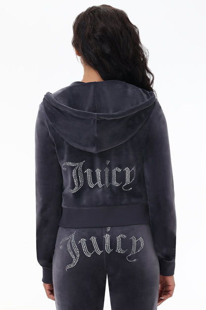 A person with long dark hair, facing away from the camera, wears the OG Big Bling Velour Hoodie—a dark gray Y2K tracksuit with a gothic "J" zipper, thumbholes, and rhinestone Juicy lettering on the back.