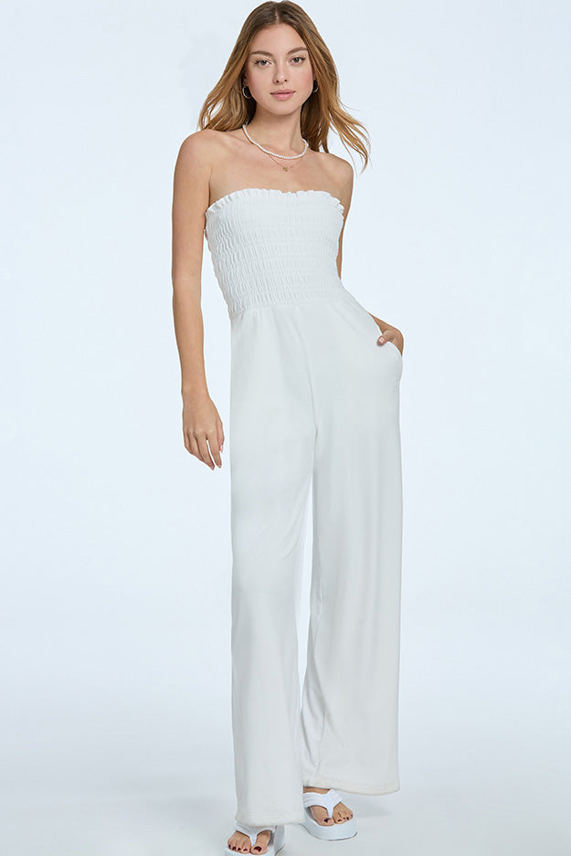 A woman with light brown hair wears the Smocked Velour Jumpsuit in white, featuring a strapless pull-on style, smocked bodice, and wide-leg pants. She pairs it with white sandals and stands against a plain light background.