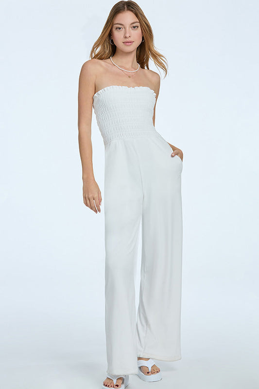 A woman with light brown hair wears the Smocked Velour Jumpsuit in white, featuring a strapless pull-on style, smocked bodice, and wide-leg pants. She pairs it with white sandals and stands against a plain light background.