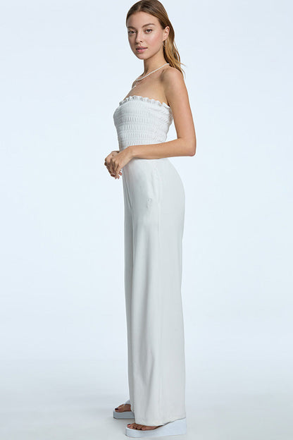 A woman in profile wears the Smocked Velour Jumpsuit—a strapless, white, smocked one-piece with wide legs. She pairs the pull-on design with light brown hair, minimal jewelry, and white platform sandals against a light background.