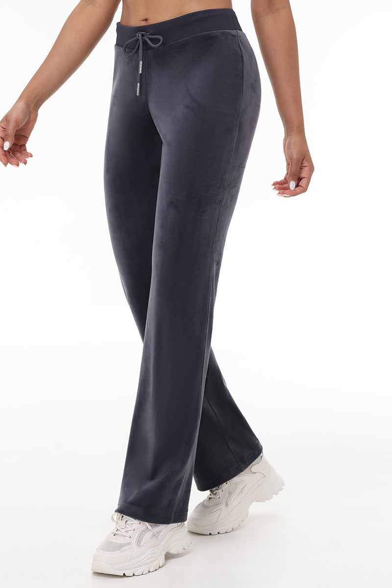 Someone models the OG Big Bling Velour Track Pants in dark gray, styled with white chunky sneakers. The photo, shot against a white background, shows only the lower half of their body, highlighting the pants’ Y2K-inspired design.