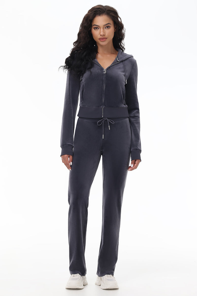 A woman faces forward in OG Big Bling Velour Track Pants and a matching dark gray zip-up hoodie with white sneakers. Her long, wavy dark hair and neutral expression channel Y2K vibes against a plain white background.