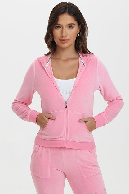 A woman with long brown hair in loose waves stands facing forward against a white background, wearing French Terry Embroidered Shorts in light pink and a matching zip-up hoodie over a white ribbed top. Her expression is neutral.