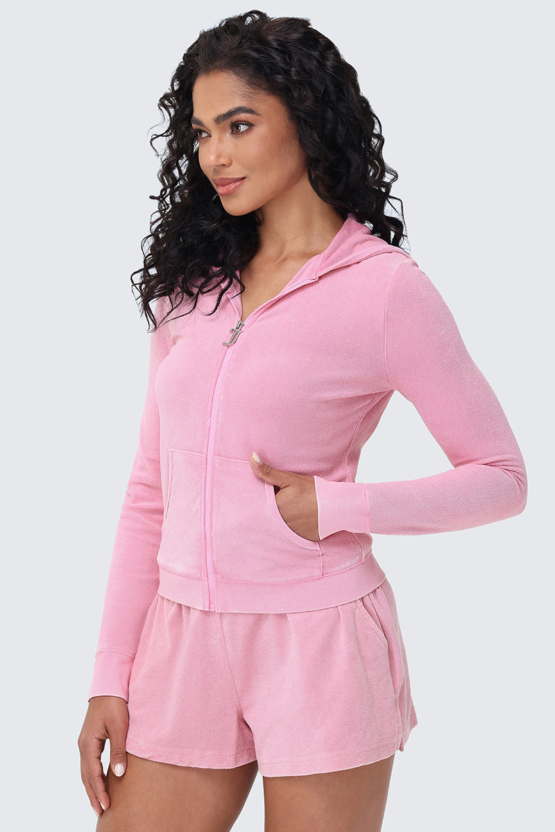 Woman with long curly hair wearing the Big Bling Washed Terry Hoodie in light pink and matching shorts, standing with one hand in her pocket and smiling slightly against a plain light background.