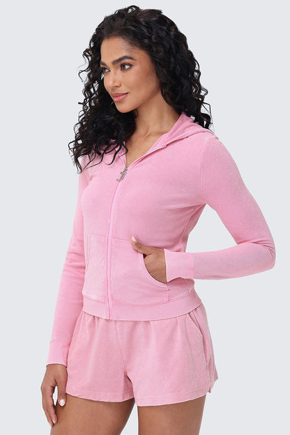 Woman with long curly hair wearing the Big Bling Washed Terry Hoodie in light pink and matching shorts, standing with one hand in her pocket and smiling slightly against a plain light background.