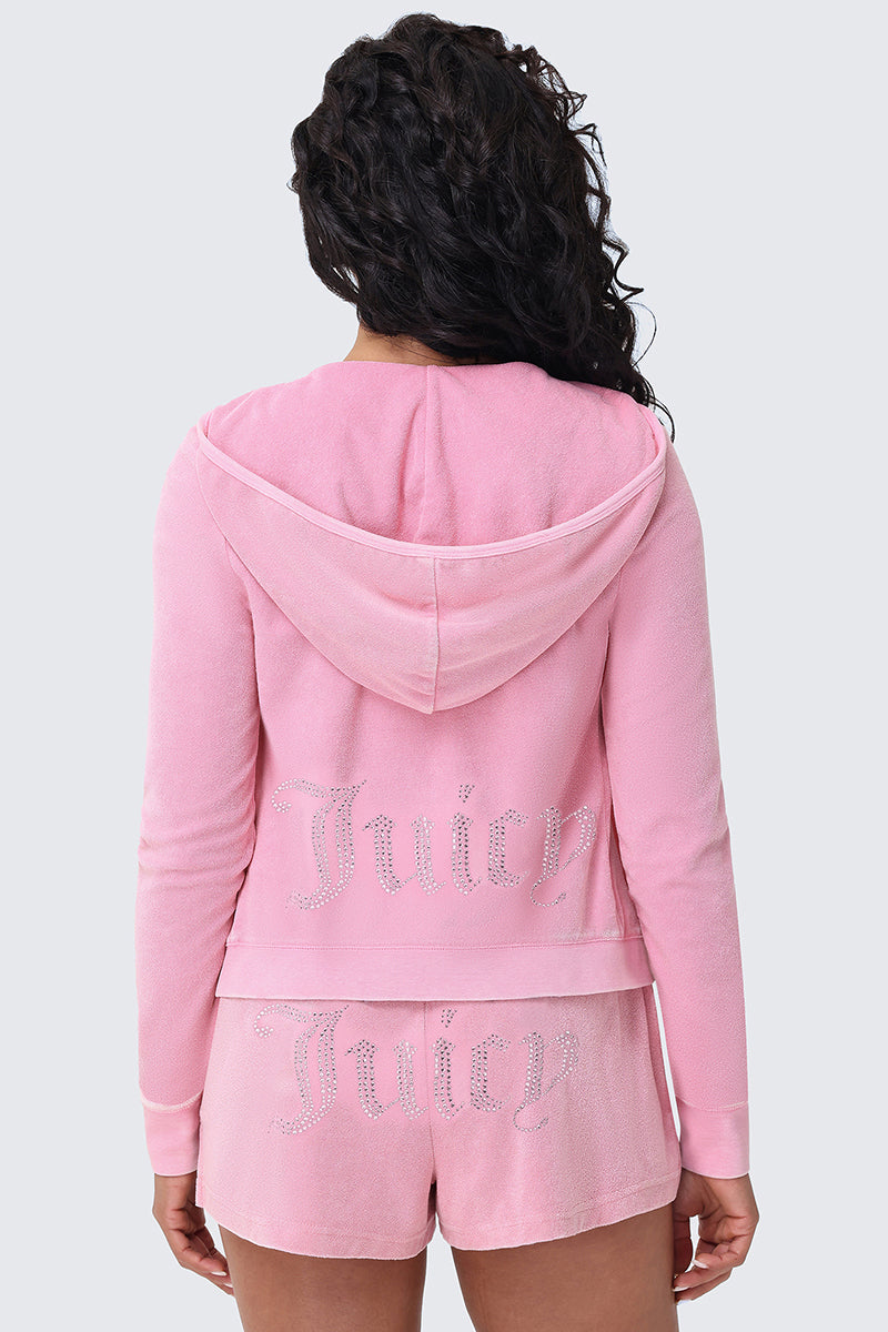 A person with long curly hair is seen from behind wearing the Big Bling Washed Terry Hoodie and matching shorts in pink, both featuring a sparkling rhinestone Juicy logo on the back and gothic J-zip detail.