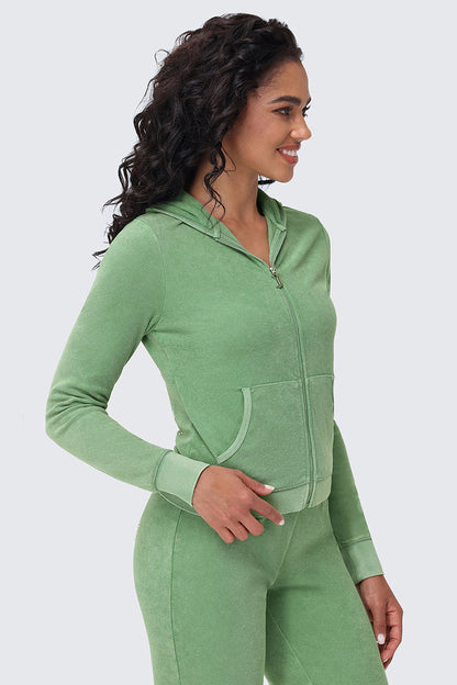 A woman with long curly hair, wearing the Big Bling Washed Terry Hoodie in light green and matching pants, stands sideways with one hand in her pocket, smiling against a plain light background.
