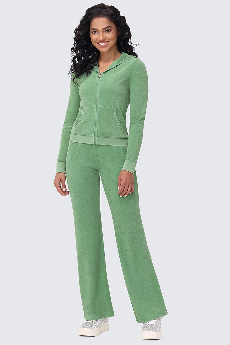 A woman with curly hair smiles while wearing the Big Bling Washed Terry Hoodie—light green, zip-up, rhinestone logo—plus matching flared pants and silver sneakers. She stands facing forward against a light gray background.