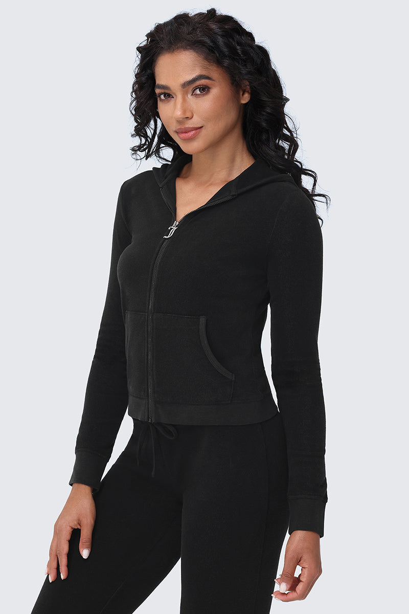 A woman with long curly hair wears the Big Bling Washed Terry Hoodie—a fitted black gothic J-zip style with front pockets—paired with matching black pants, standing against a plain light gray background.