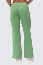 A person wearing Big Bling Washed Terry Track Pants in green, featuring a rhinestone "Juicy" logo across the back, stands hands on hips and pairs them with white platform shoes.