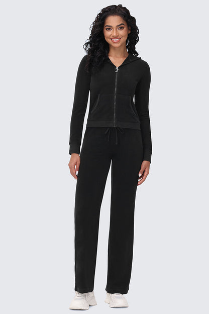 A woman faces forward, smiling, in Big Bling Washed Terry Track Pants with a black zip-up hoodie and white sneakers, bringing beach-chic style to a plain light gray background.
