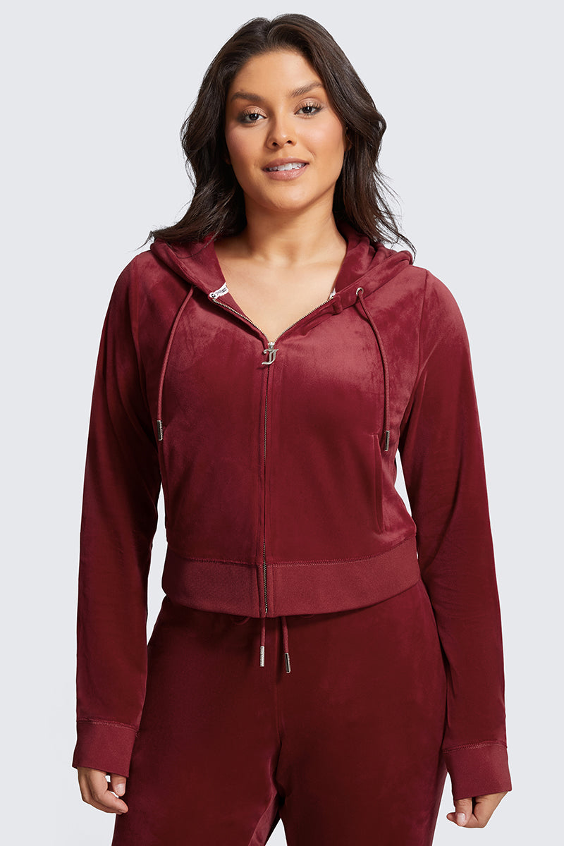 A woman wears the Plus-Size OG Big Bling Velour Hoodie in maroon, zipped up with a gothic "J" zipper. She faces forward, smiling slightly, her dark hair loose over her shoulders against a plain light gray background.