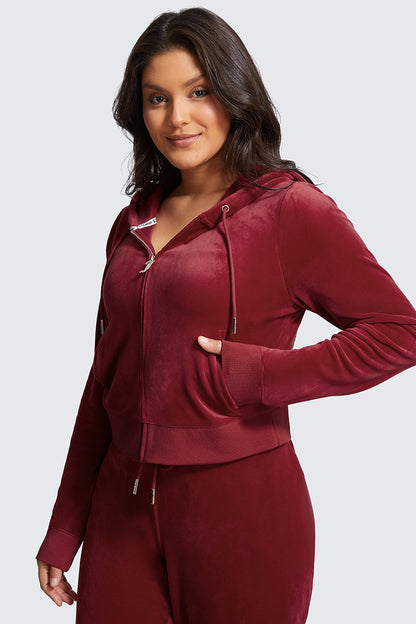 A woman with long dark hair models the Plus-Size OG Big Bling Velour Hoodie in burgundy with thumbholes, paired with matching pants, standing against a light gray background and smiling gently at the camera.